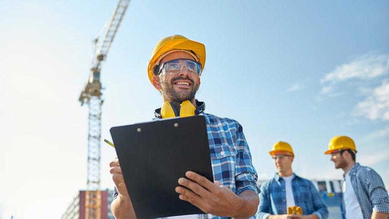 Three workers stand at a construction site on a sunny day. One man is in the foreground and two are standing in the background. They are each wearing a yellow hardhat and a blue shirt over a white t-shirt. The man in front is holding a clipboard.